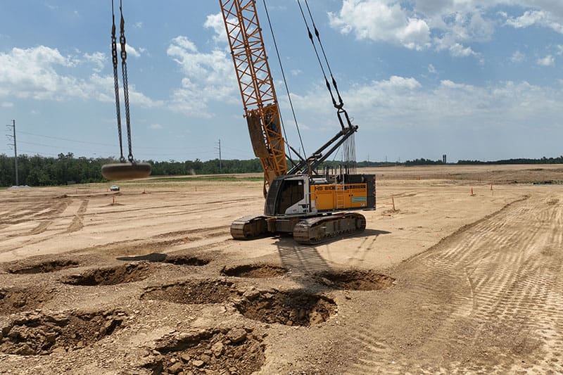 Crawler crane dropping a heavy weight over cratered soil during dynamic compaction on a construction site.