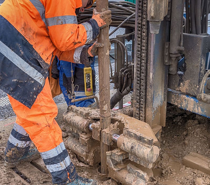 worker-operating-drilling-equipment-ground-densification Worker operating geotechnical drilling equipment to test soil conditions on a muddy construction site during ground investigation.