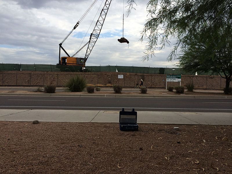 roadside-dynamic-compaction-with-monitoring-equipment-densification Crawler crane lifting tamper weight for dynamic compaction while vibration monitoring equipment records ground movement near roadway.