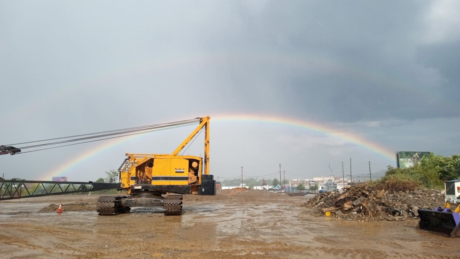 crawler-crane-on-muddy-site-under-double-rainbow-densification A yellow crawler crane on a muddy construction site with a double rainbow arching across a grey sky in the background.