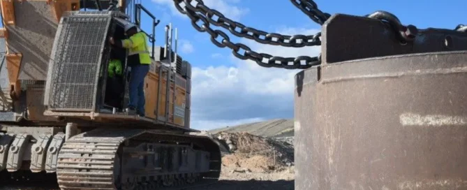 A close-up of a construction worker in a high-visibility vest standing on the tracks of a large crane, with heavy metal chains connected to a massive steel weight in the foreground.
