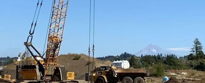 A large crawler crane with a lattice boom standing on a dirt construction site next to a yellow dump truck, with a snow-capped mountain visible in the distant background.