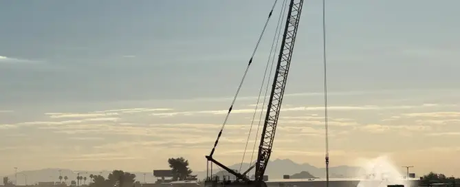 Crawler crane operating over a dynamic compaction site at dusk with a water truck suppressing dust nearby