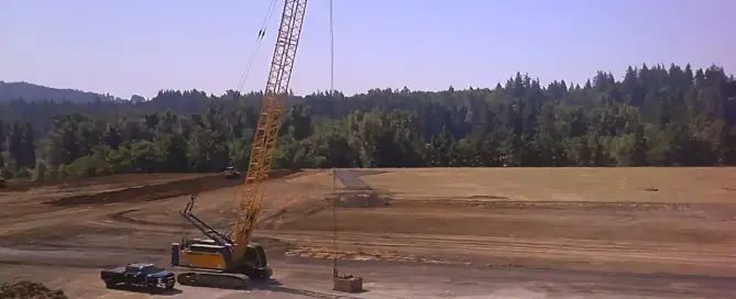Crawler crane raising a dynamic compaction weight over a graded construction site near a forest