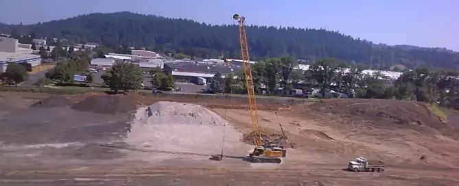 Dynamic compaction crane lifting a tamping weight above a soil stockpile at an industrial site