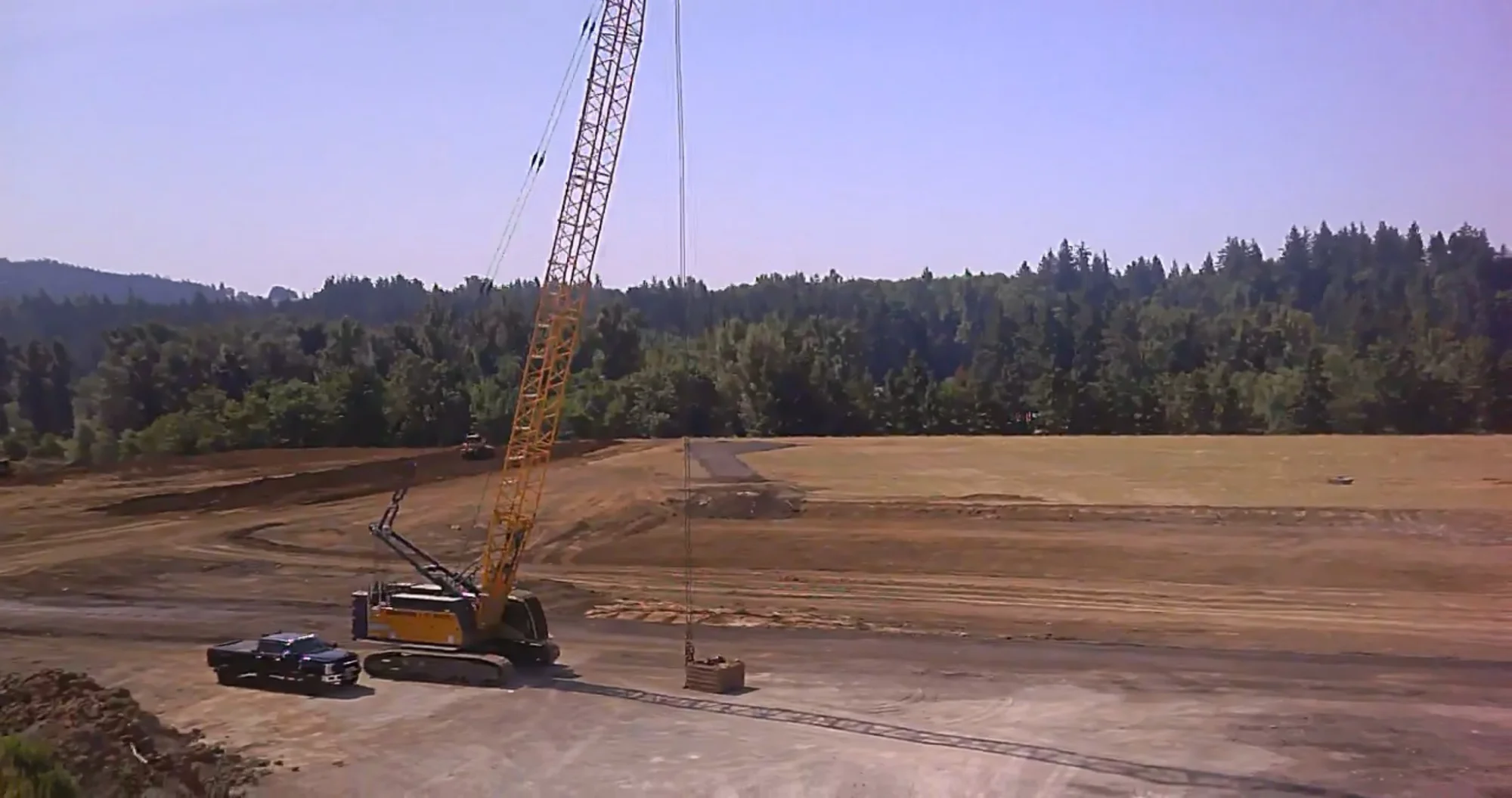crawler-crane-dynamic-compaction-site-densification. Crawler crane raising a dynamic compaction weight over a graded construction site near a forest